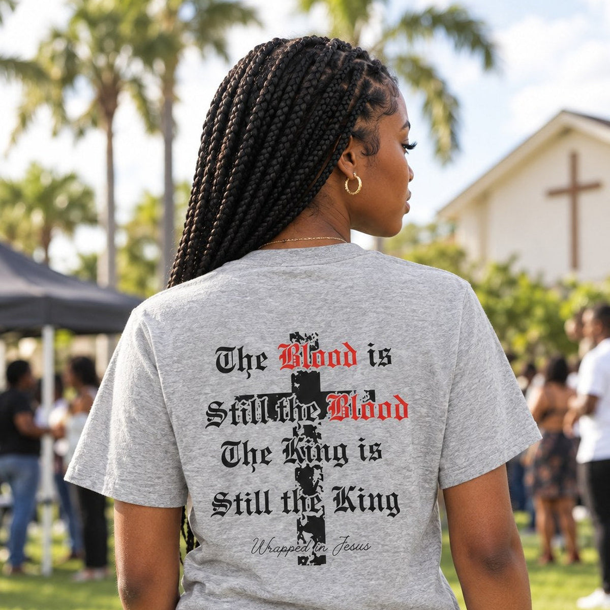 Woman wearing heather gray The Blood Is Still the Blood Christian tee at an outdoor church gathering, back graphic view