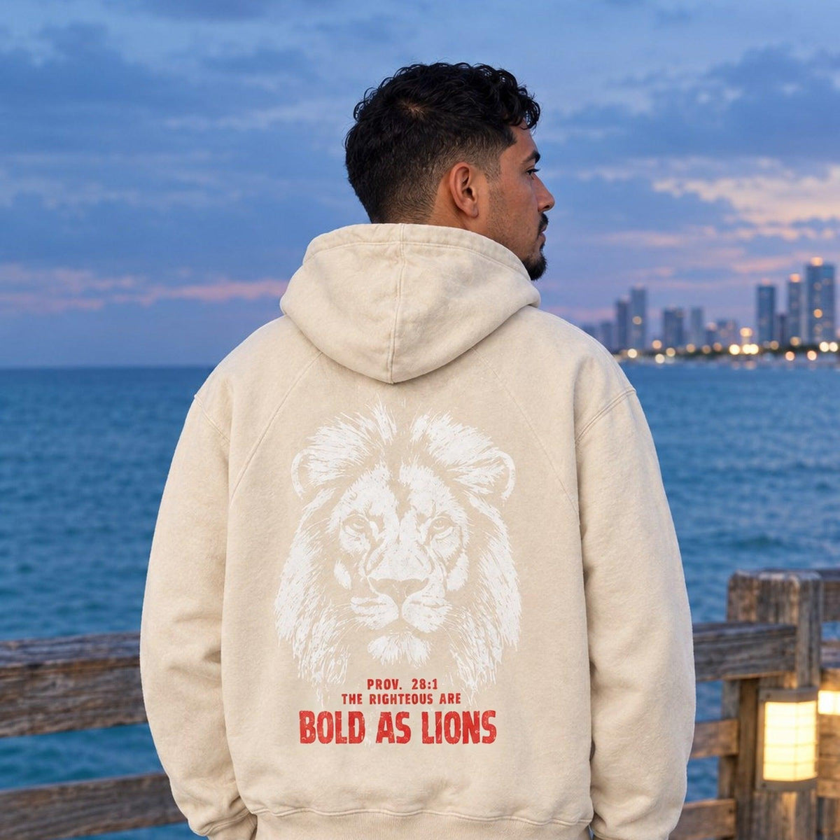 Man wearing cream Eternal hoodie with Bold as Lions lion back graphic on a Miami Beach pier at blue hour